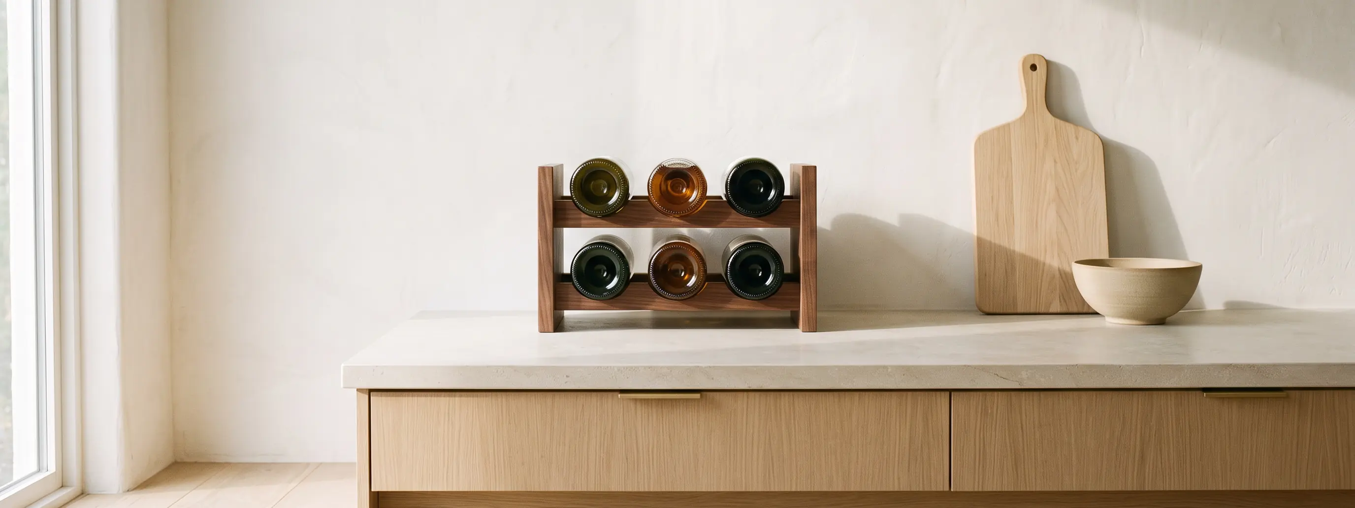 A small walnut countertop rack holding six bottles on their sides on a pale limestone counter, an oak board and a ceramic bowl beside it